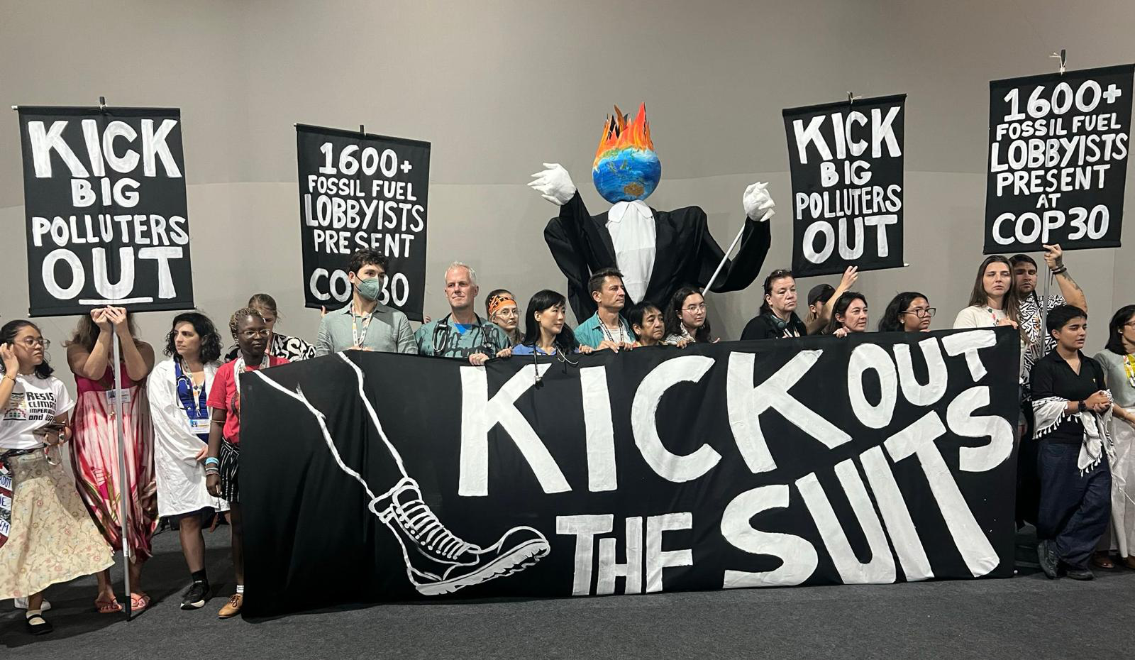 CAPE team, partners and supporters holding a large banner at a COP30 event: 'Kick Out the Suits', and smaller signs: 'Kick Big Polluters Out' and '1600+ Fossil Fuel Lobbyists Present at COP30', alongside a structure in a shape of a corporate executive with a globe on fire as a head